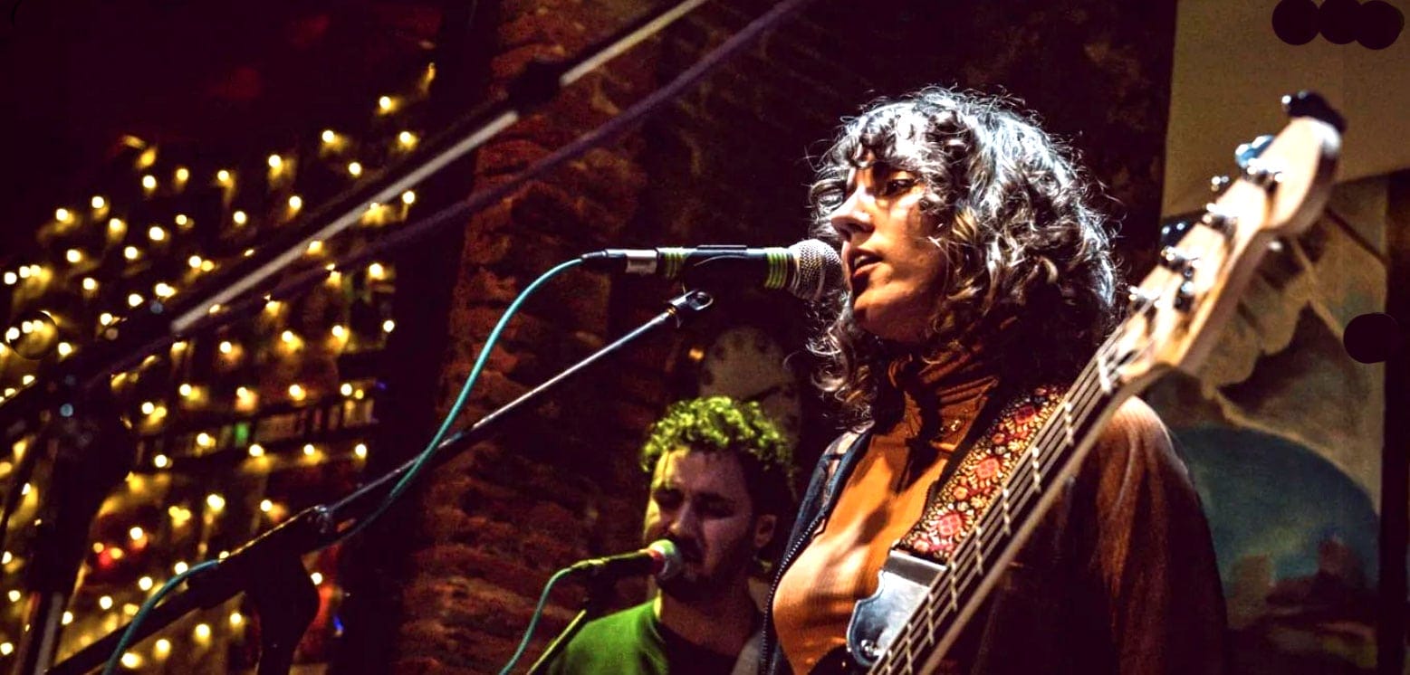 Live music performance featuring a female singer with a guitar at a cozy, intimate venue with warm lighting and decorative string lights.