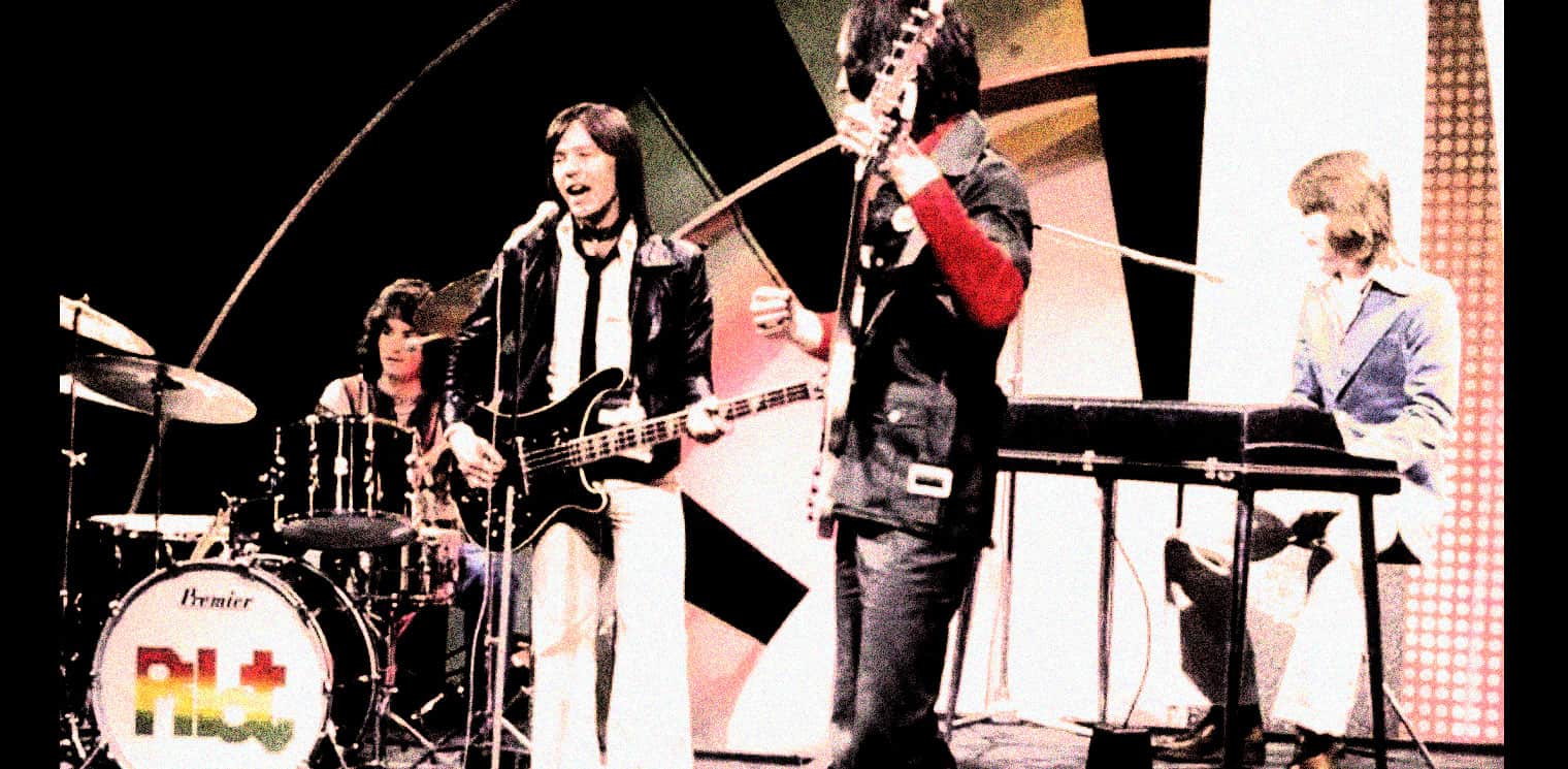 Vintage rock band performing live on stage, featuring guitar, drums, and keyboard, celebrating classic music history.
