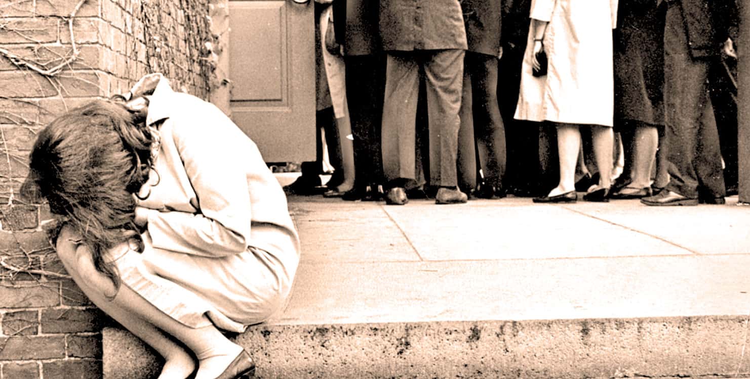 Girl sitting alone on sidewalk, appears distressed or homeless, surrounded by a group of people. Image evokes themes of social issues, homelessness, and urban life, capturing a moment of solitude amid a crowd.