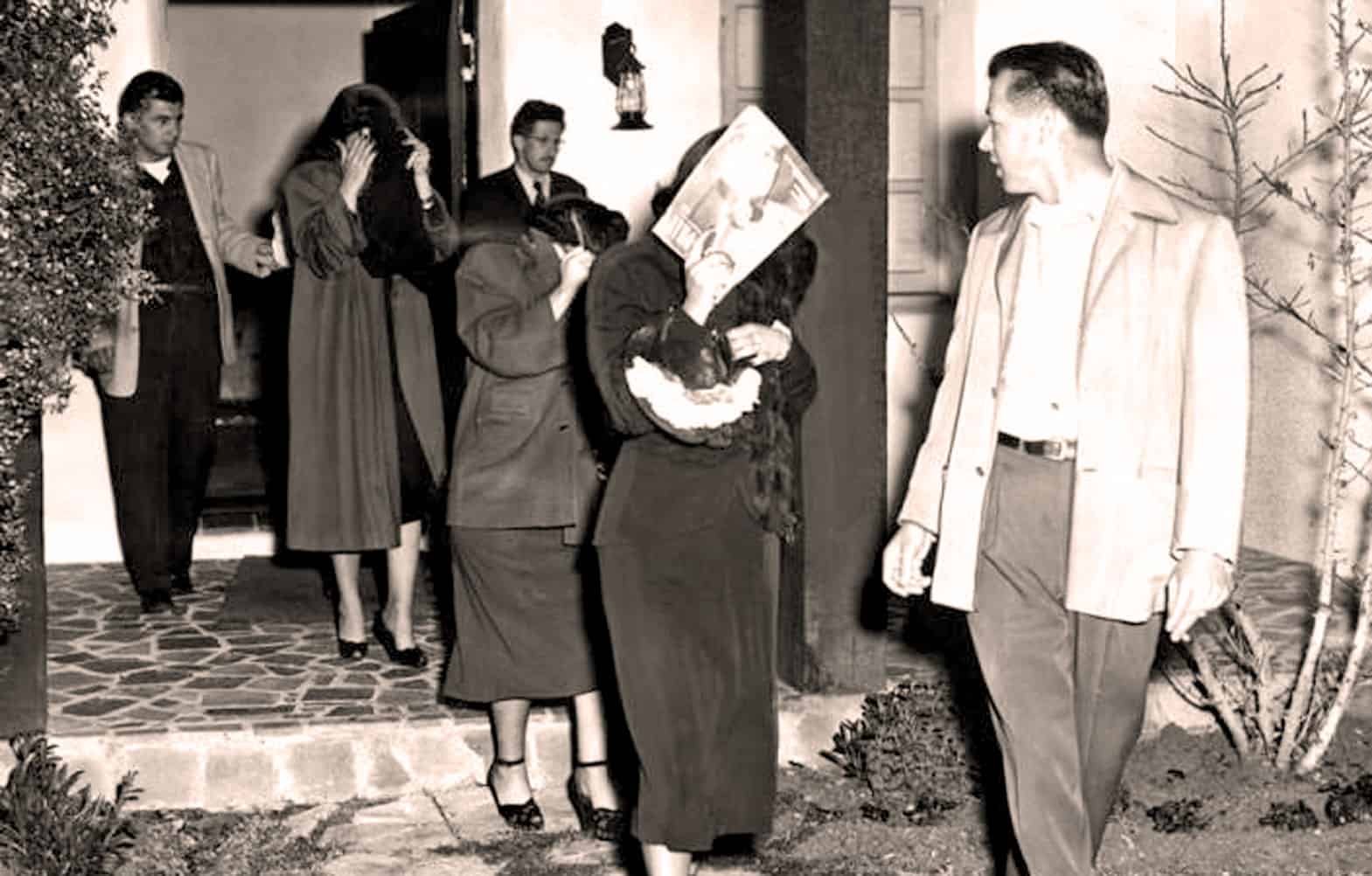 People standing in line outside a building, some covering their faces, with a man and woman talking nearby; vintage black and white photo offering historical context for news and cultural archives.