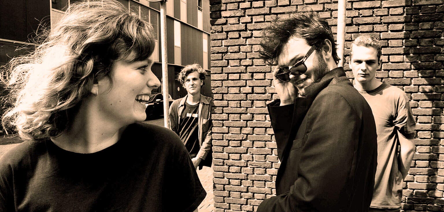 Smiling young woman talking with man wearing sunglasses and black jacket, urban street scene, friendship, casual conversation, modern city environment, storytelling, social interaction, diverse group of friends, candid moment, black and white photo.