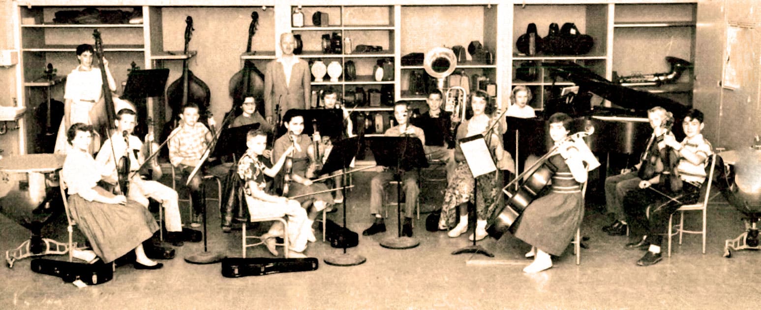 Vintage children's orchestra practicing with string instruments in a music classroom, featuring music stands and various musical instruments on shelves, capturing a nostalgic educational and musical era.