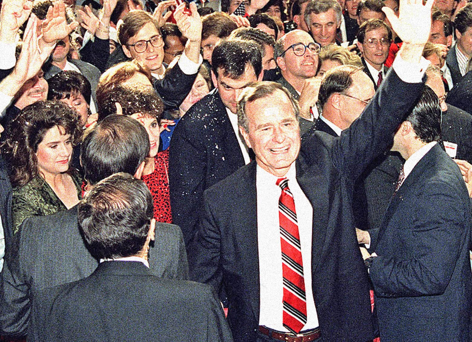 A lively political rally featuring a man in a suit and red-striped tie waving to a crowded audience, surrounded by supporters and media, capturing a significant moment in news and history coverage.