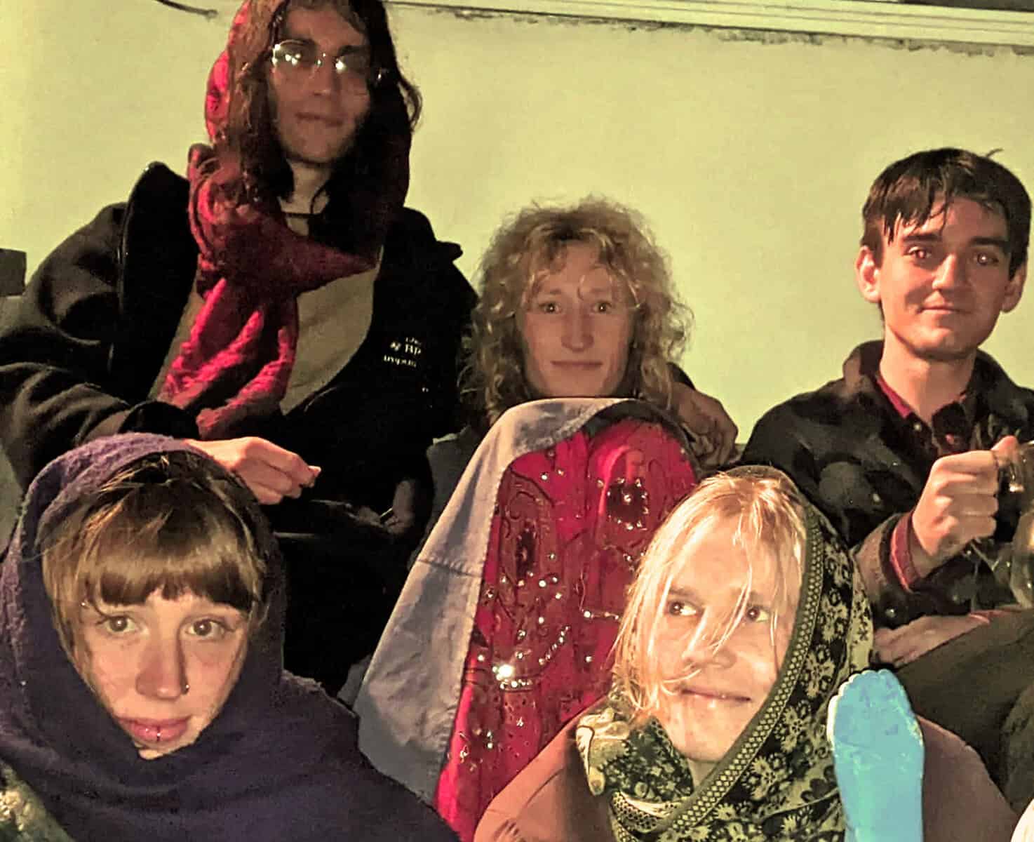 People gathered in a room, some wearing colorful scarves, with a background related to music, news, or history archives, highlighting a vintage or nostalgic atmosphere.