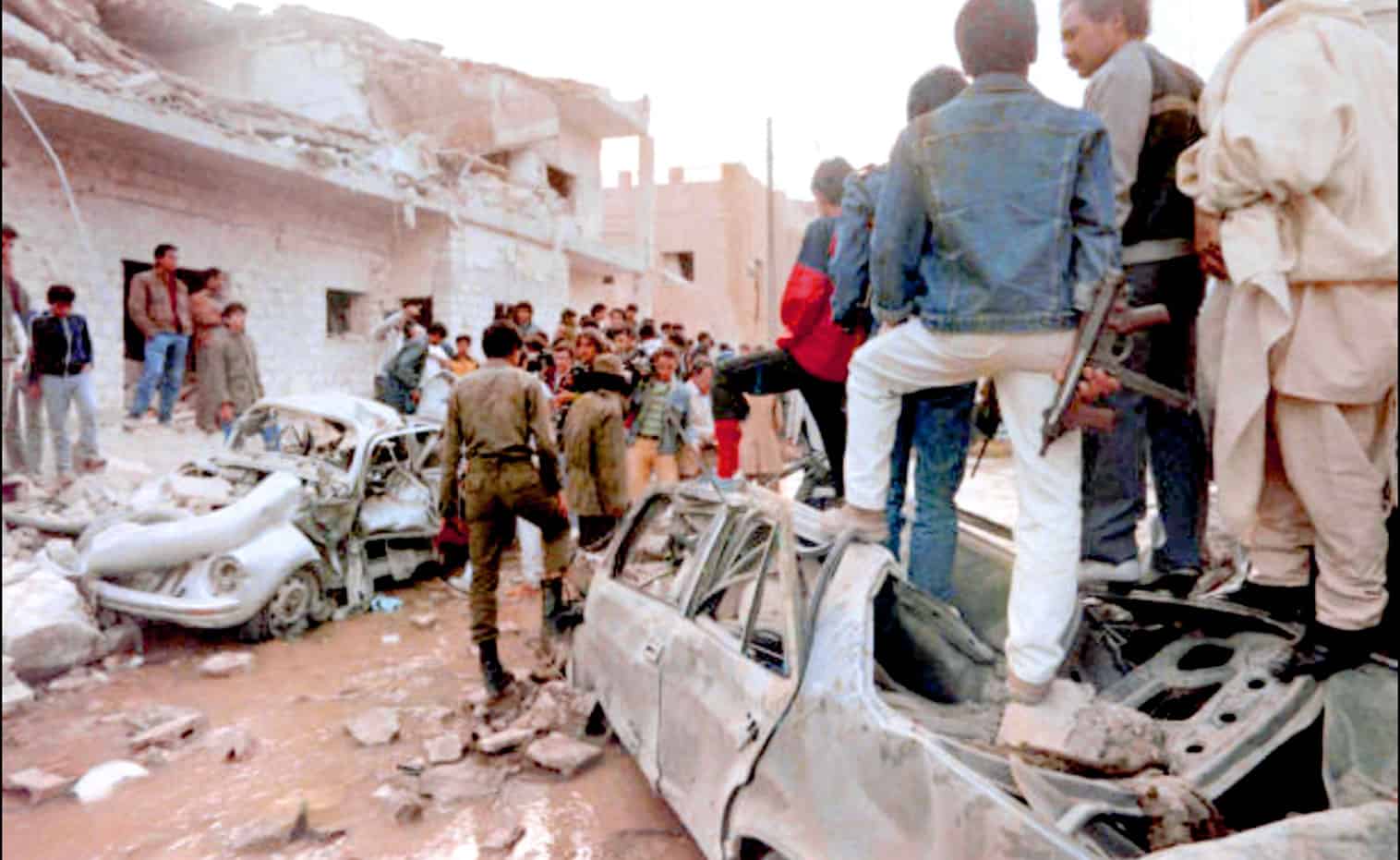 Destroyed cars and a crowd of people in a war-torn urban area.