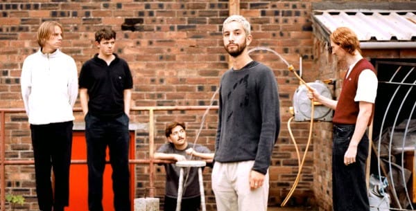 A group of young adults standing outdoors against a brick wall, engaging in conversation with vintage radio equipment; nostalgic scene related to music and radio history.