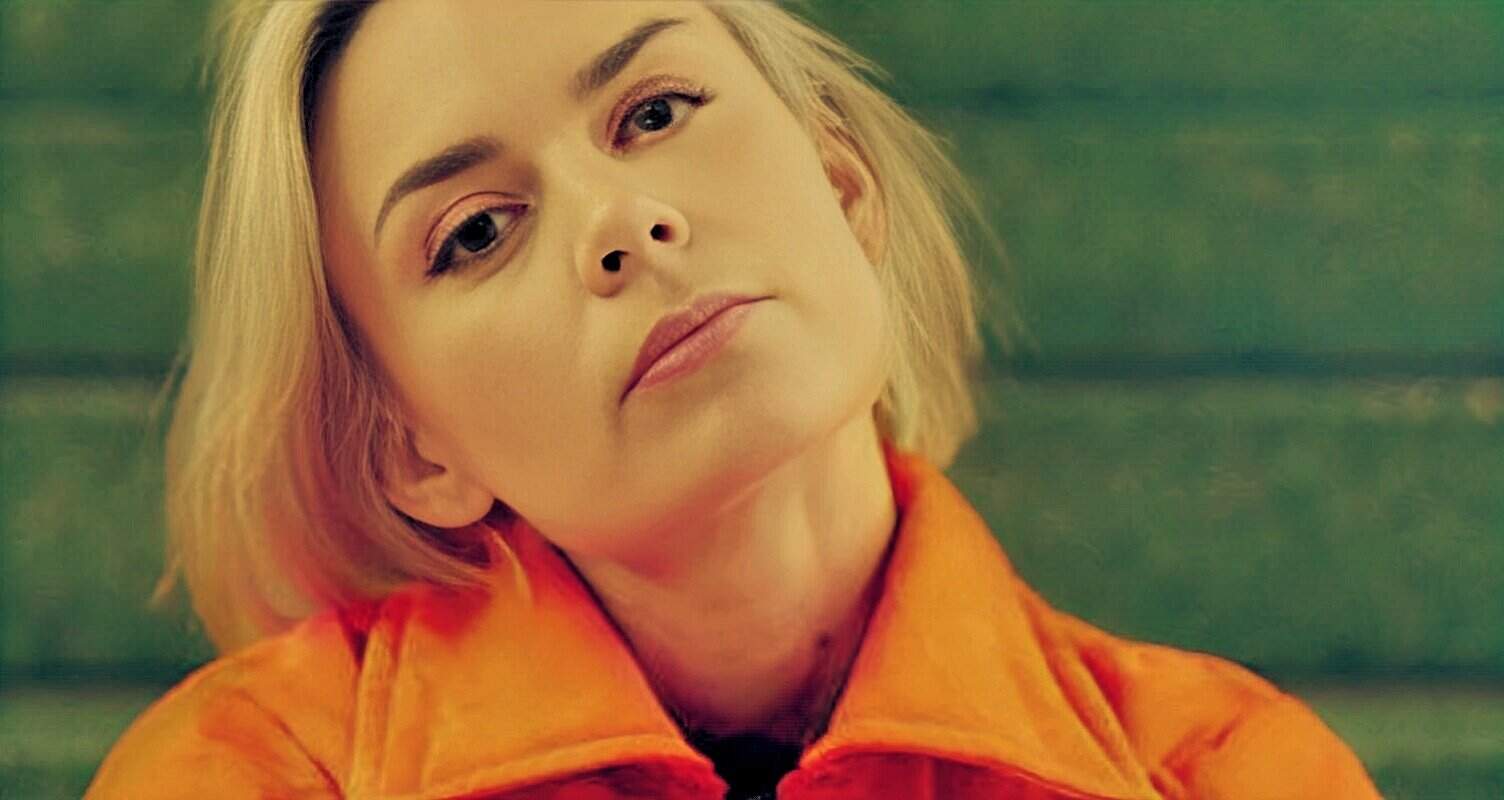 Woman with short blonde hair and orange jacket looking at camera, vibrant background, artistic portrait for music or news history content.