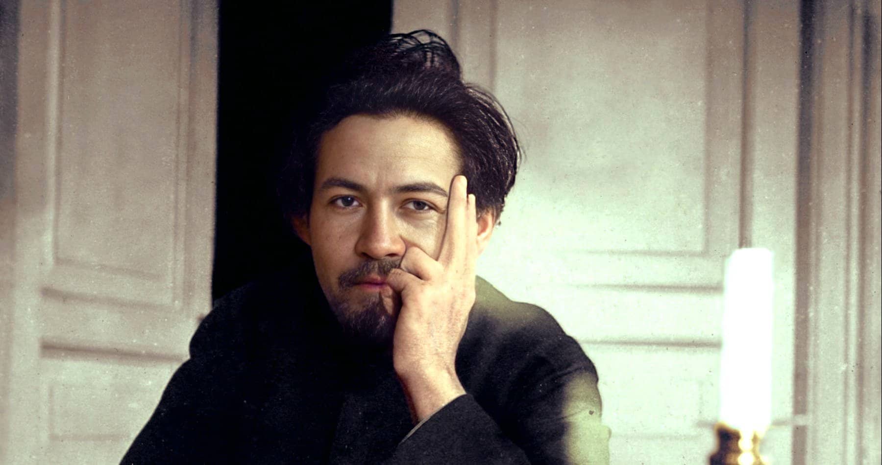 Portrait of a man with tousled dark hair and a contemplative expression, sitting at a table with a lit candle, in a classic indoor setting reflecting music, history, and news archival themes.