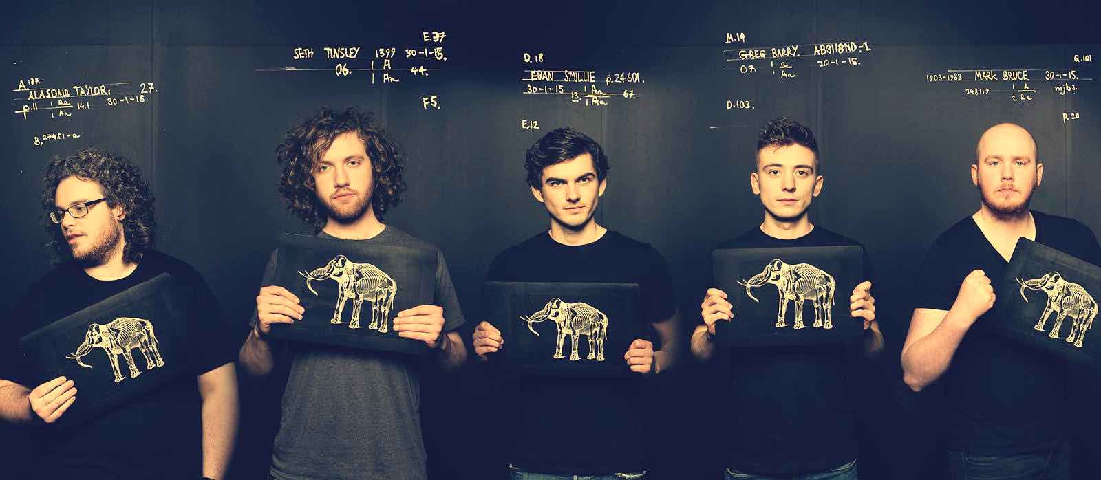 Elephant graphic on black folders held by five young men, music band members, in front of a blackboard with handwritten music notes and dates, showcasing a creative music or band concept.