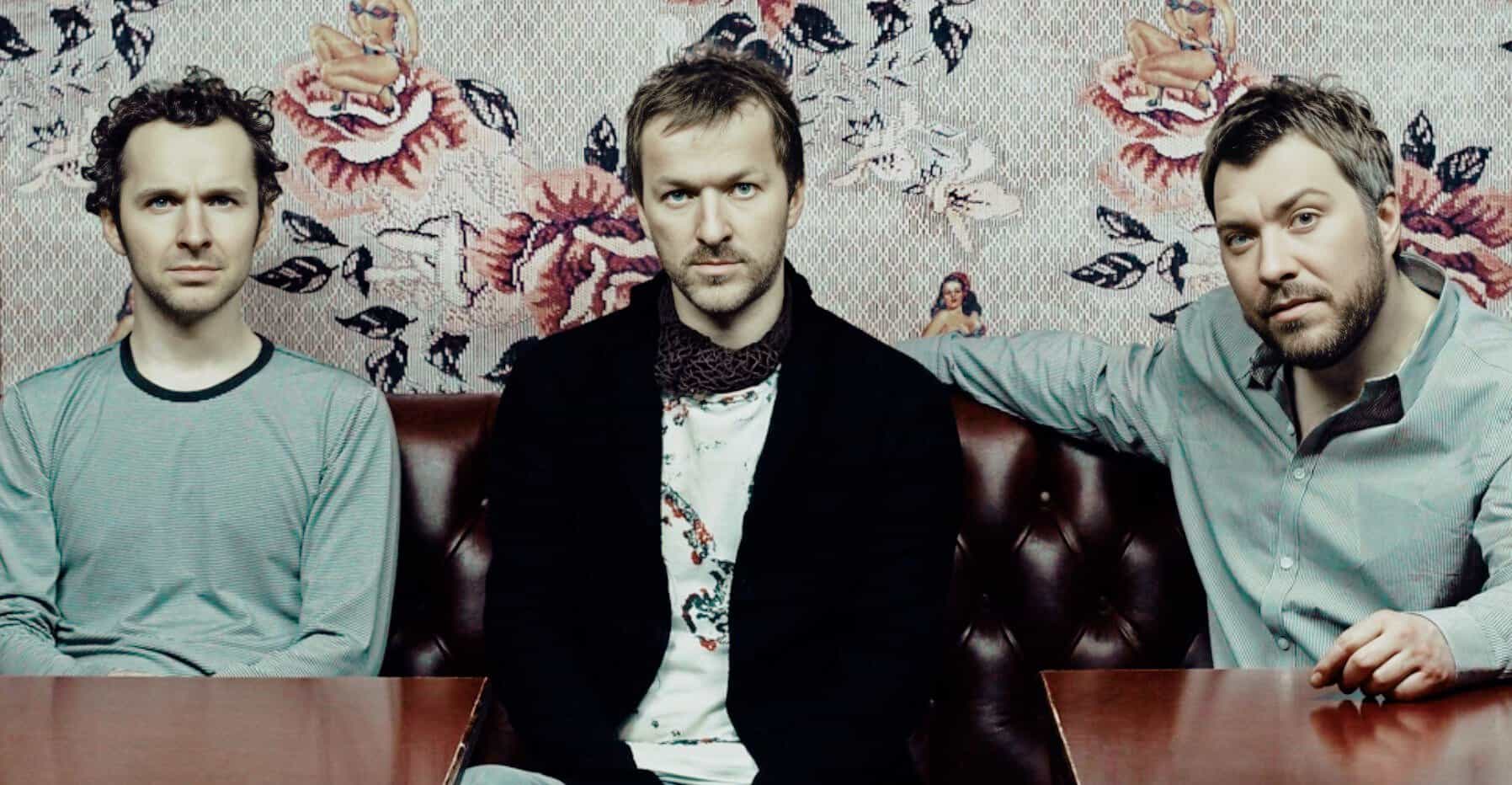 Artistic photo of three men sitting on a vintage sofa with floral wallpaper background, alternative rock band members, modern music and news archive photography, Past Daily source relevance.
