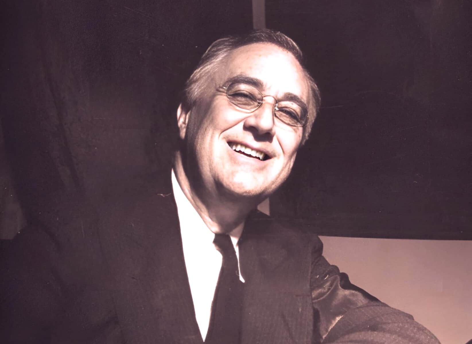 Vintage black-and-white portrait of a smiling man in a suit and glasses, representing historical news, music, and cultural archives available at Past Daily.
