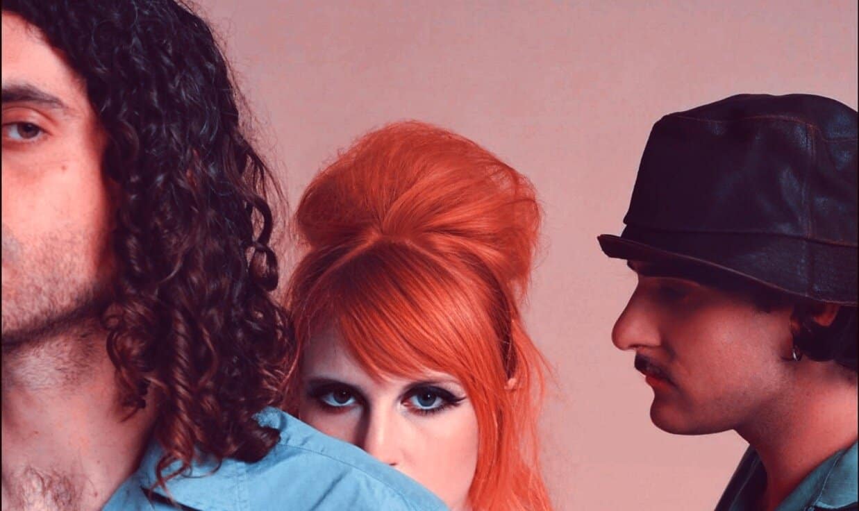 Bold red-haired woman with dramatic makeup and styled hair, flanked by two men, one with long curly hair and the other with a black leather cap, in a vibrant, edgy photo for music or news archives.