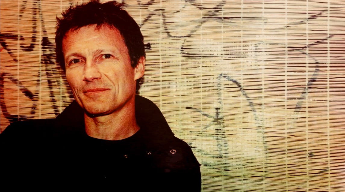 Rick Rubin, music producer, in front of bamboo wall with graffiti, showcasing the artistic vibe of the Past Daily sound archive of news, history, and music.