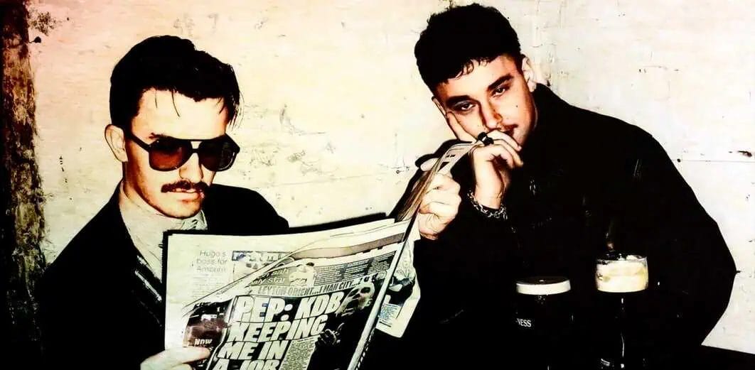 Pepe Kobre and a friend enjoying drinks in a vintage setting, with one reading a newspaper and the other holding pints of dark beer, capturing a retro musical vibe.