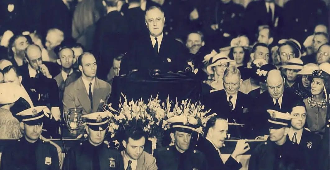 A historical black-and-white photograph of Franklin D. Roosevelt delivering a speech at a formal event, surrounded by an attentive audience of men and women, many wearing hats, during the era of the New Deal.