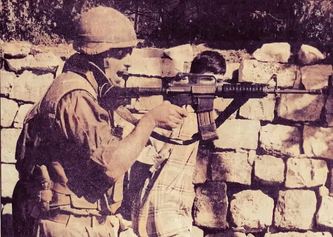 Faded black and white image of a soldier aiming a rifle with a scope during a military operation, positioned against a stone wall with trees in the background.
