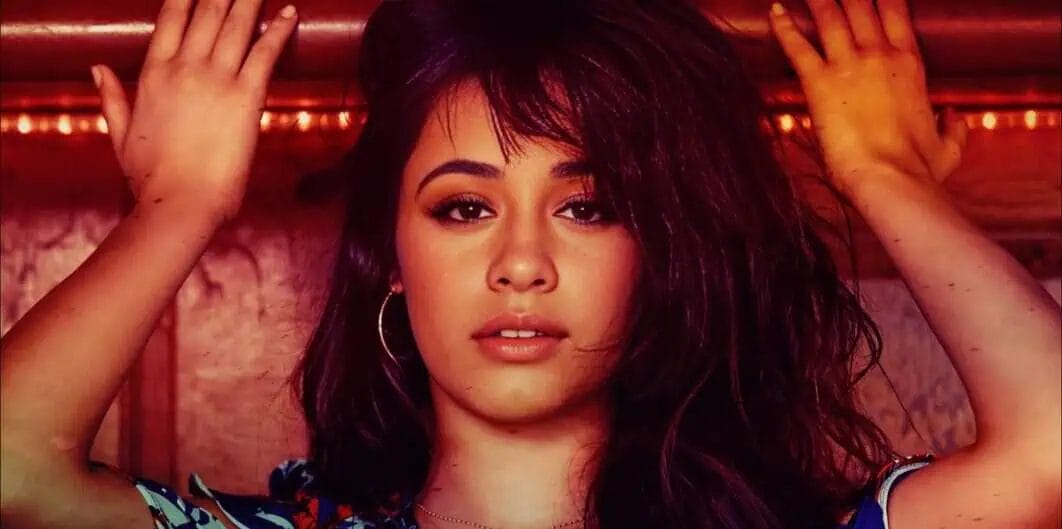 Women with long dark hair posing indoors, head tilted, hands raised near her head, wearing hoop earrings, vibrant lighting and textured background.