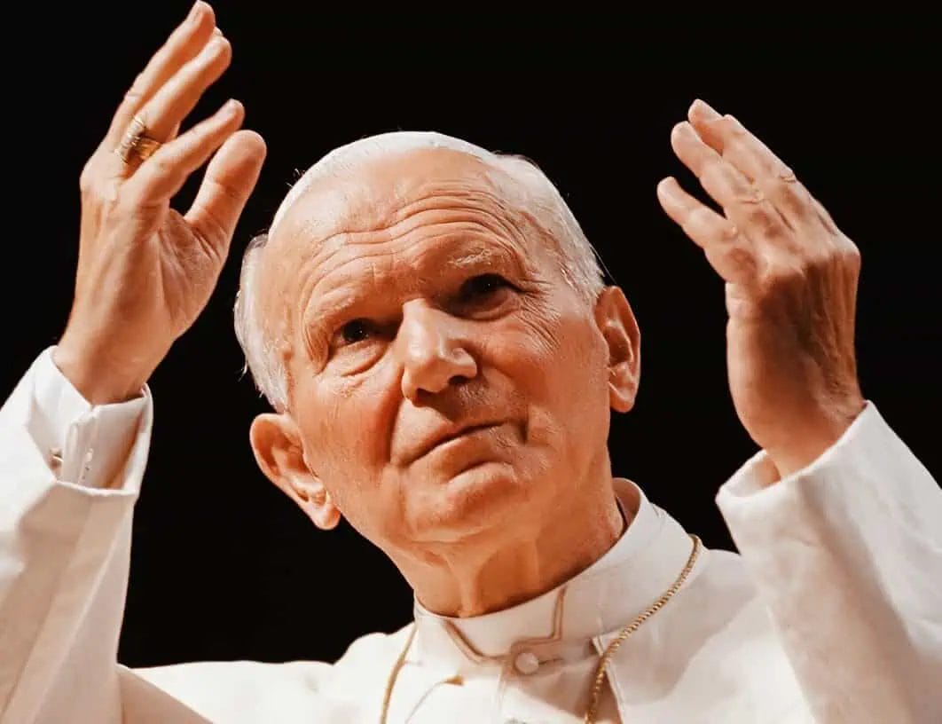 Elderly man with white hair making a gesture with both hands, wearing a white robe and gold necklace.